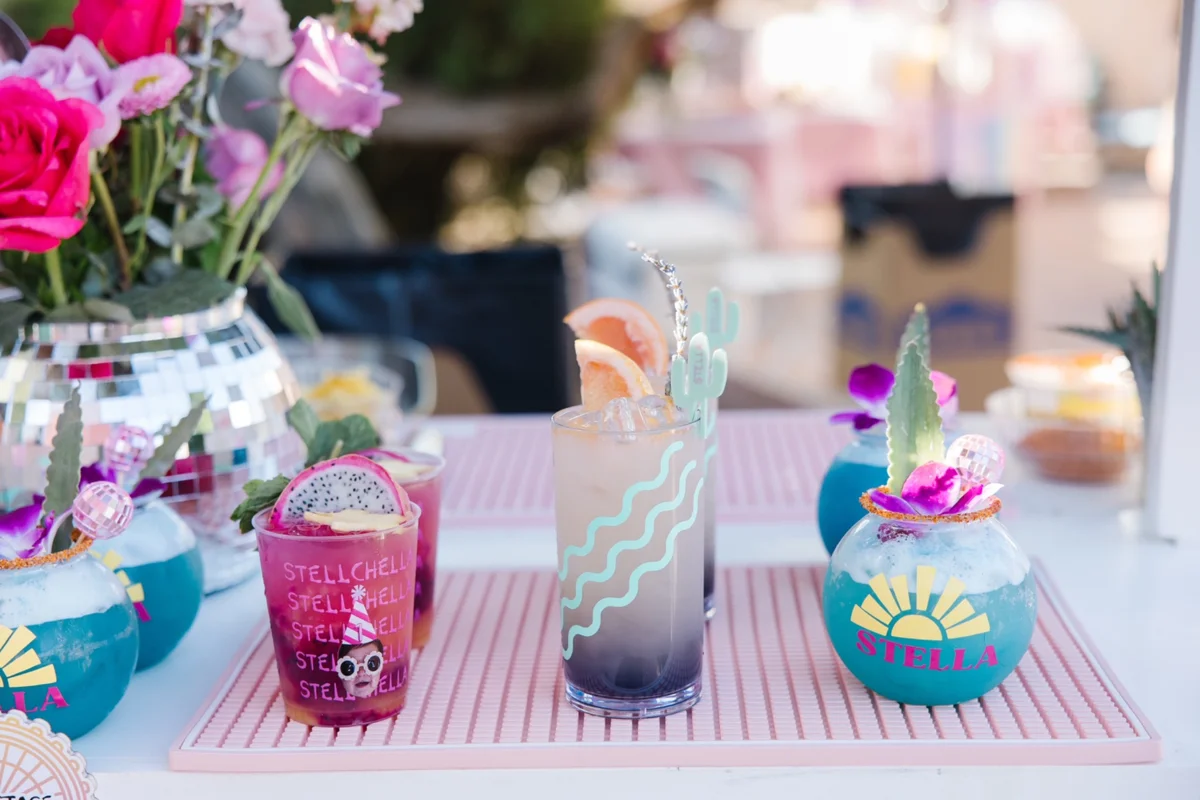 Three colorful signature cocktails on a pink bar with flowers and a custom menu